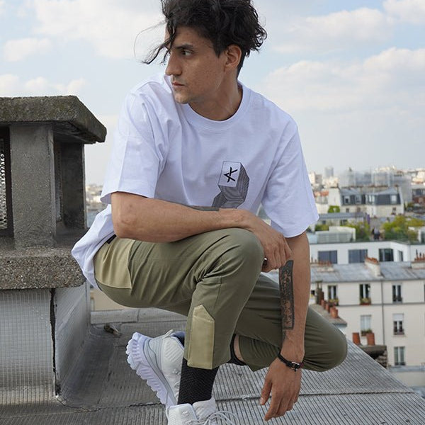 Homme accroupi en extérieur portant un pantalon Taitsu kaki, coupe droite ajustée à la cheville, avec empiècements contrastés et fermeture magnétique